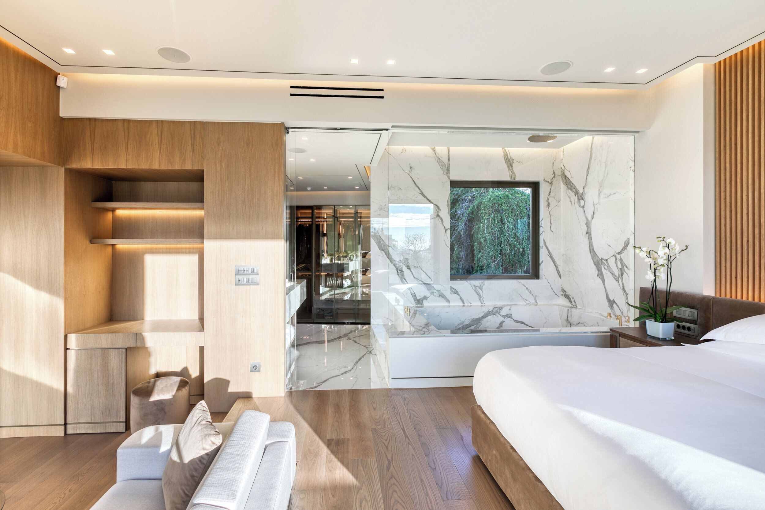 Een moderne hotelkamer met een groot bed, een zithoek met een bank en een kleine tafel, een accentmuur van marmer, en uitzicht op het omliggende landschap.