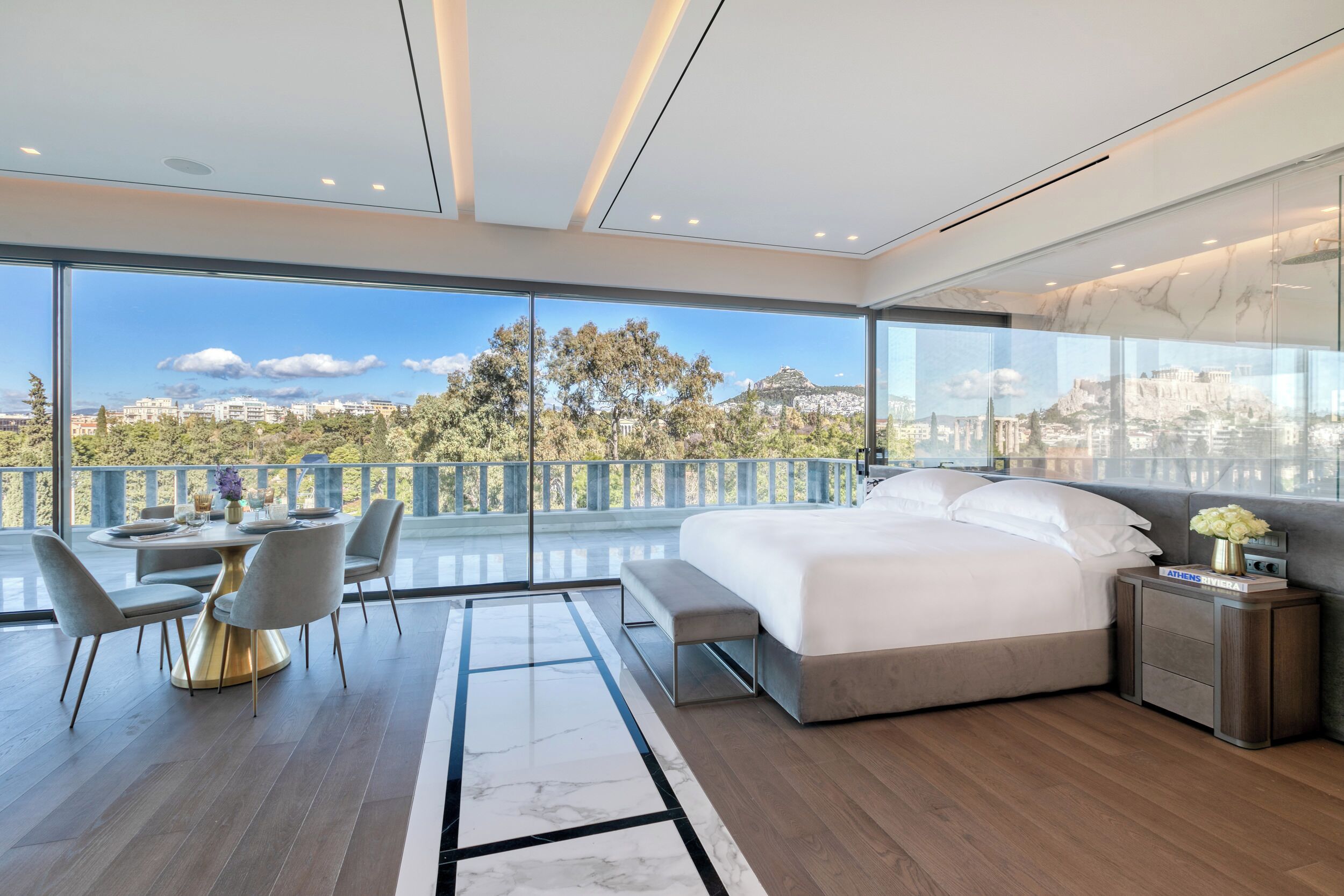 Quarto moderno com uma cama grande, área de refeições com mesa redonda e cadeiras, e varanda com vista para a cidade.