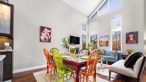Een eetruimte met een tafel voor vier personen, omringd door kleurrijke stoelen. Er hangen schilderijen aan de muur, een televisie op een standaard en een bankstel op de achtergrond.