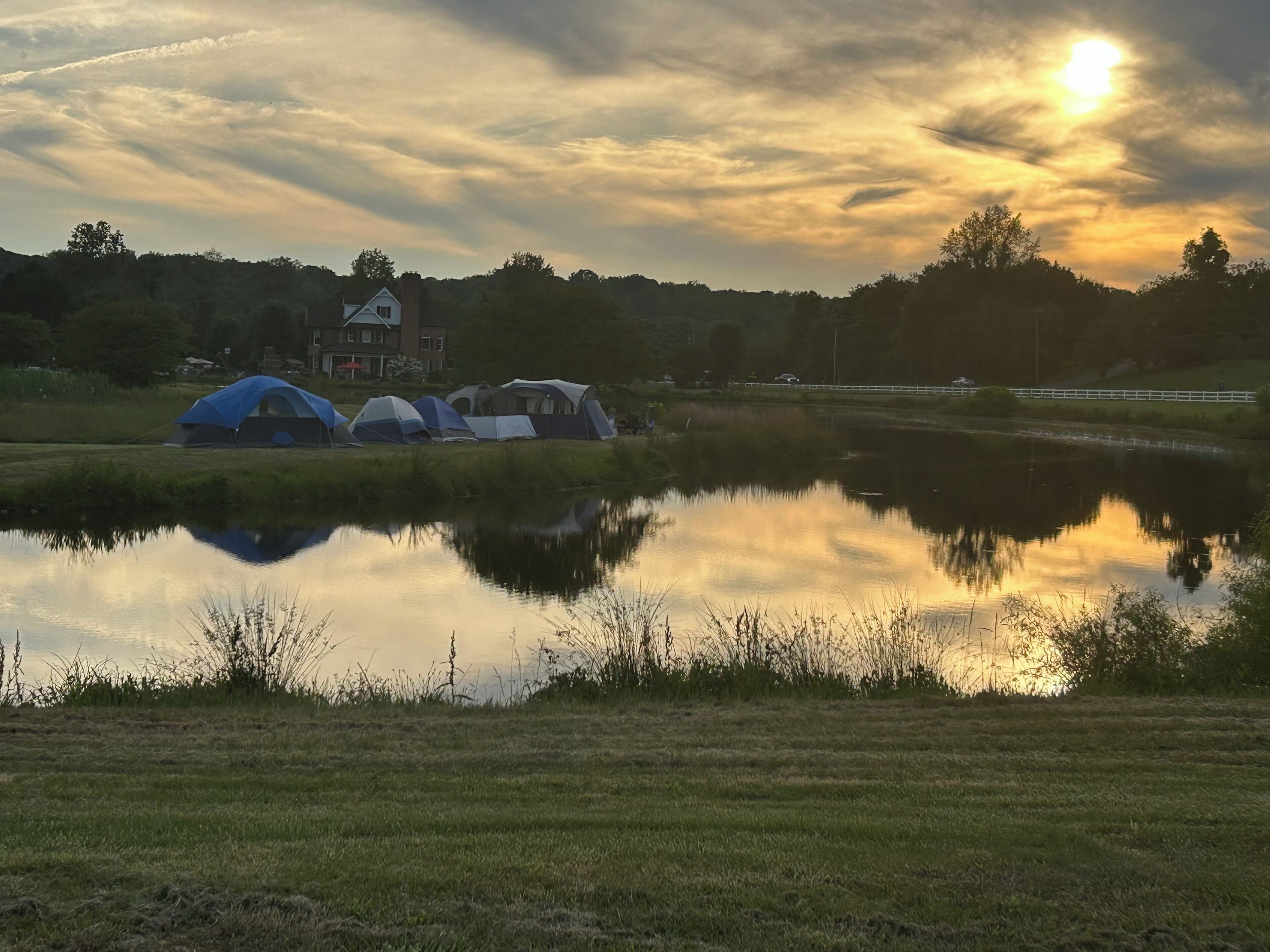 A campsite with tents near a body of water, a house in the background, and a sunset.