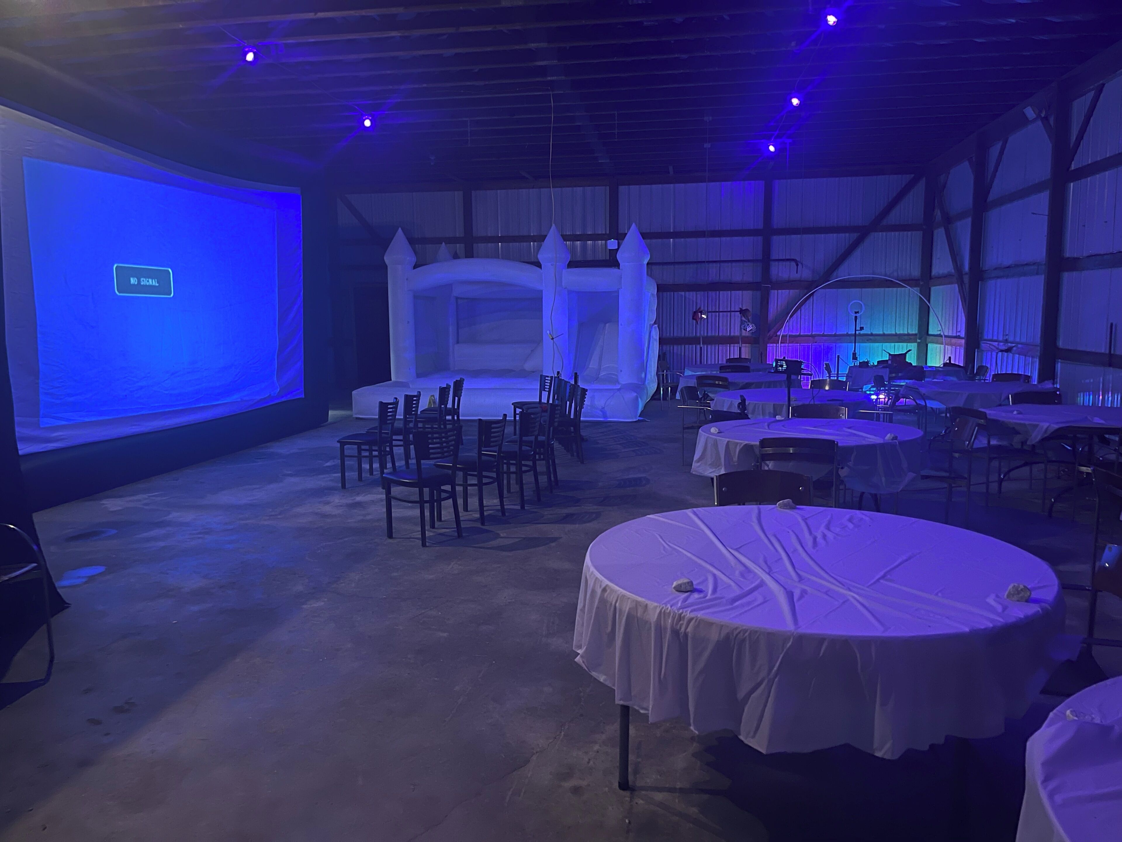 A banquet hall with round tables covered in white tablecloths, a stage with a blue screen, and a backdrop with a 'NO SIGNAL' message.