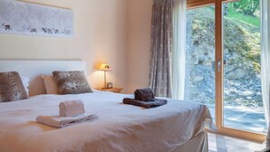Habitación de hotel con una cama grande, una mesita de noche con una lámpara y vistas a una zona rocosa al exterior a través de una puerta corrediza de vidrio.