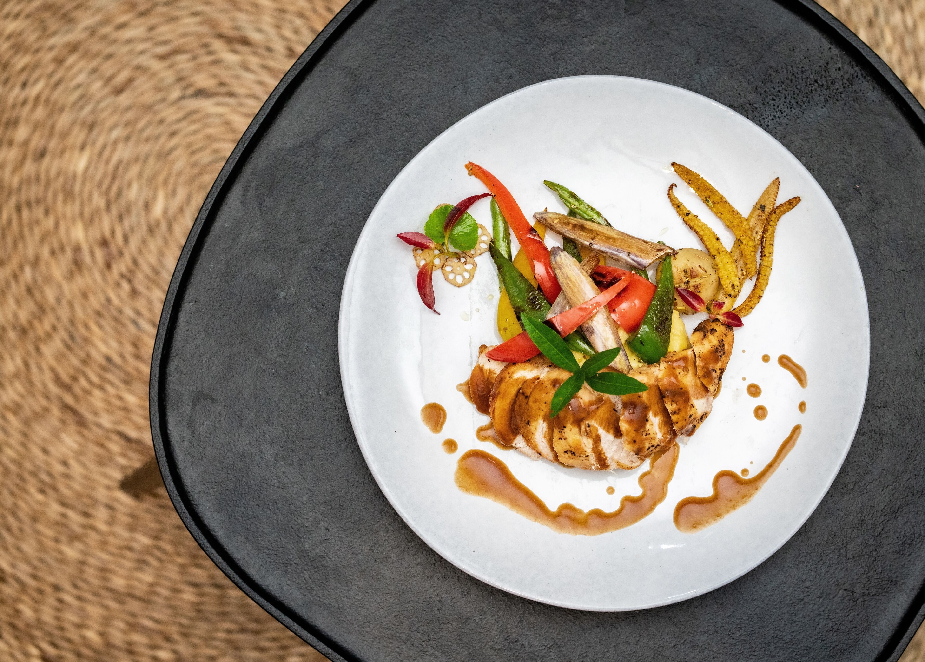 Un plato blanco con pollo a la parrilla, verduras y salsa sobre un plato oscuro.