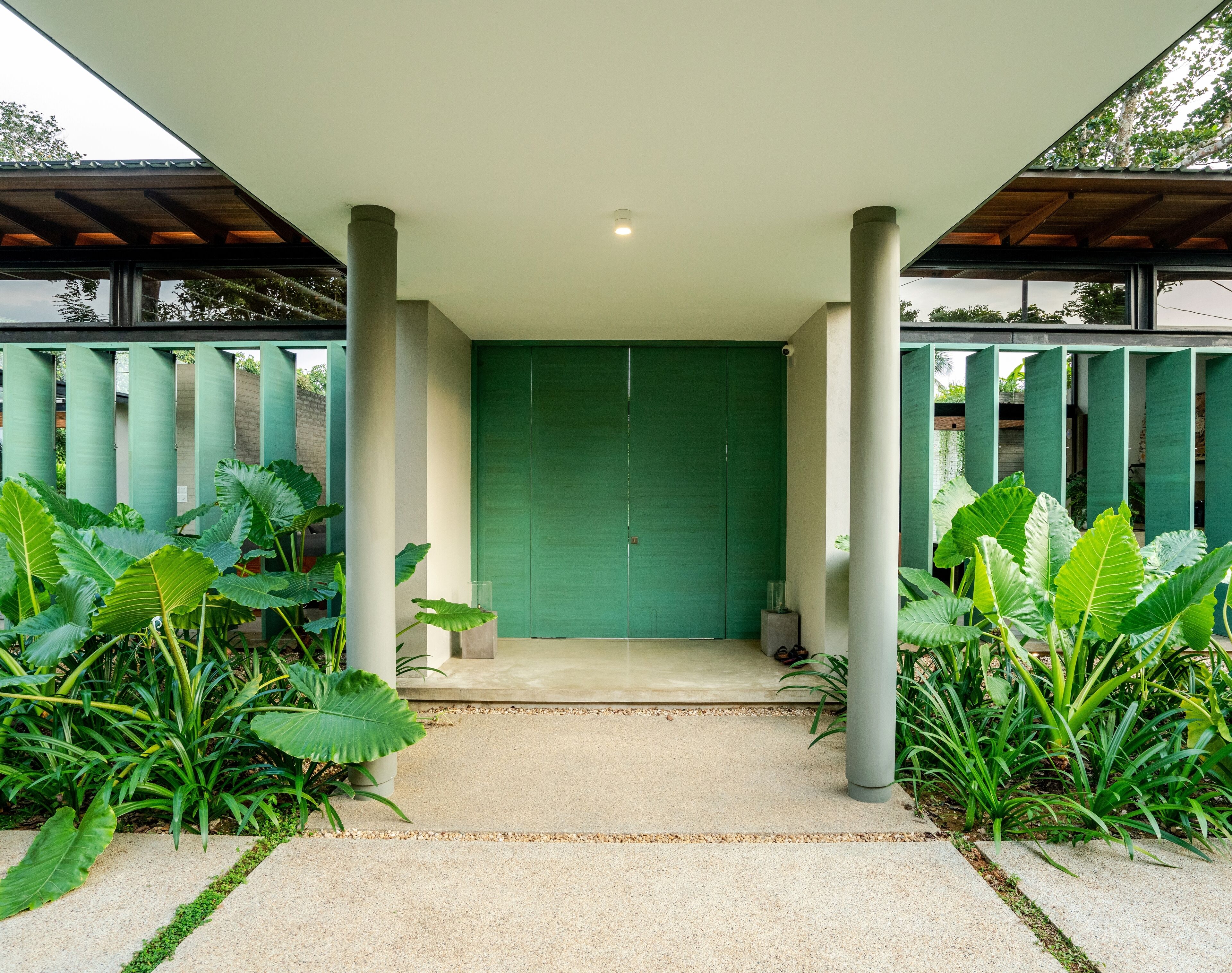 A covered entrance with a green door, surrounded by large plants and columns.