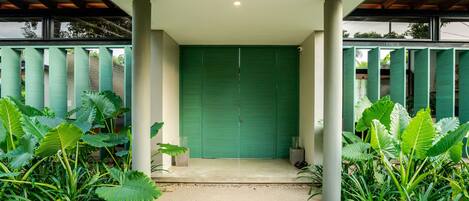 A covered entrance with a green door, surrounded by large plants and columns.