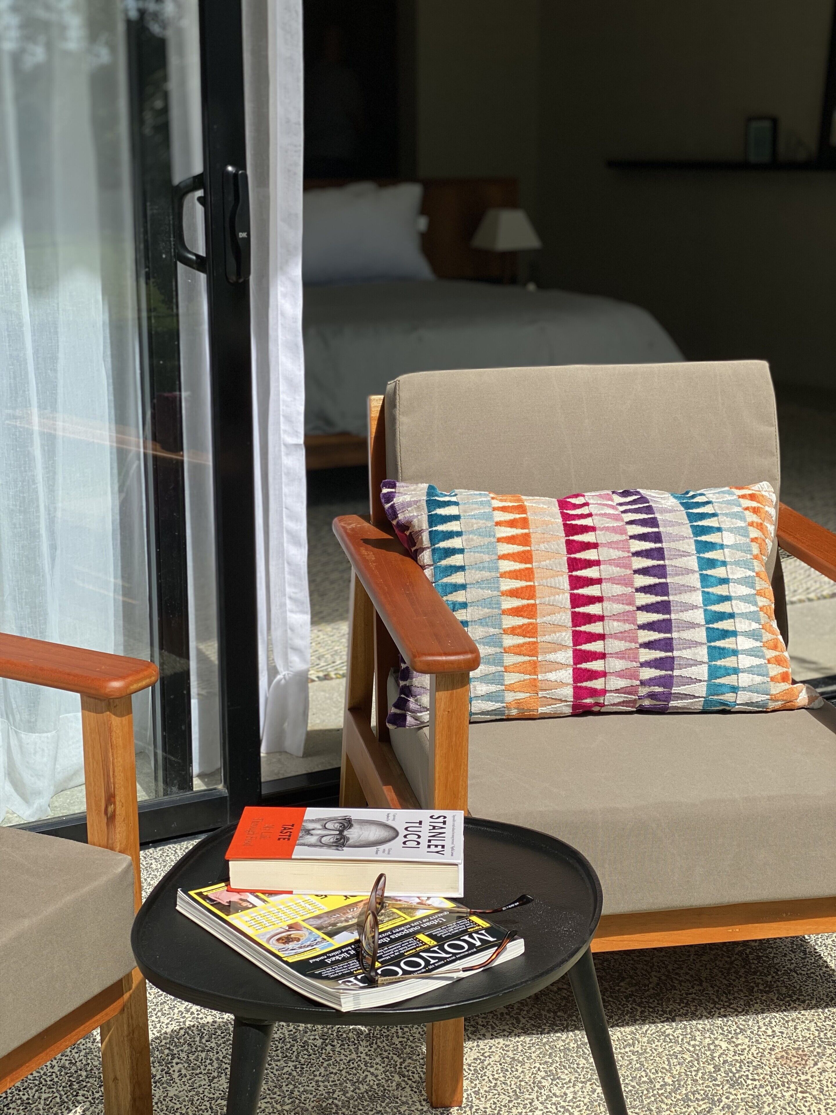A hotel room with a beige armchair, a colorful pillow, a small table with a magazine and glasses, and a bed with a white pillow.