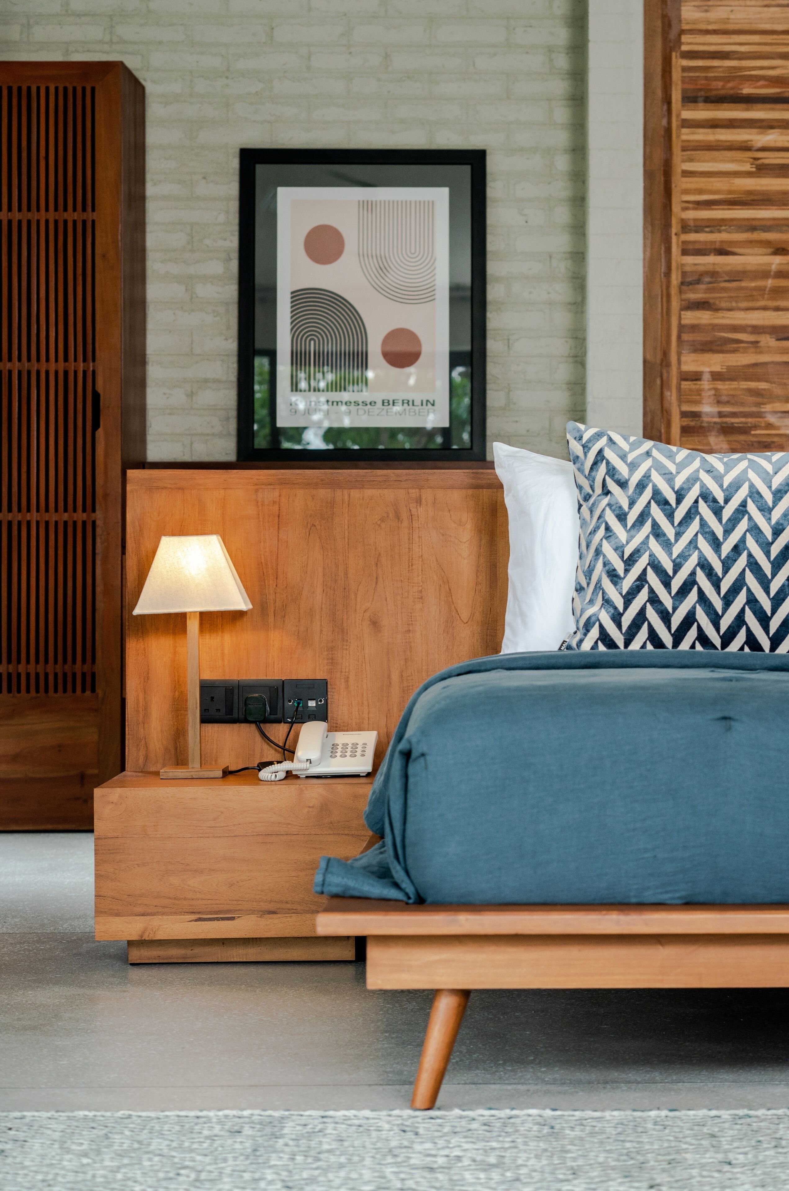 Een moderne slaapkamer met een houten hoofdbord, een nachtkastje met een lamp, een ingelijste afbeelding en een blauw beddegoed.