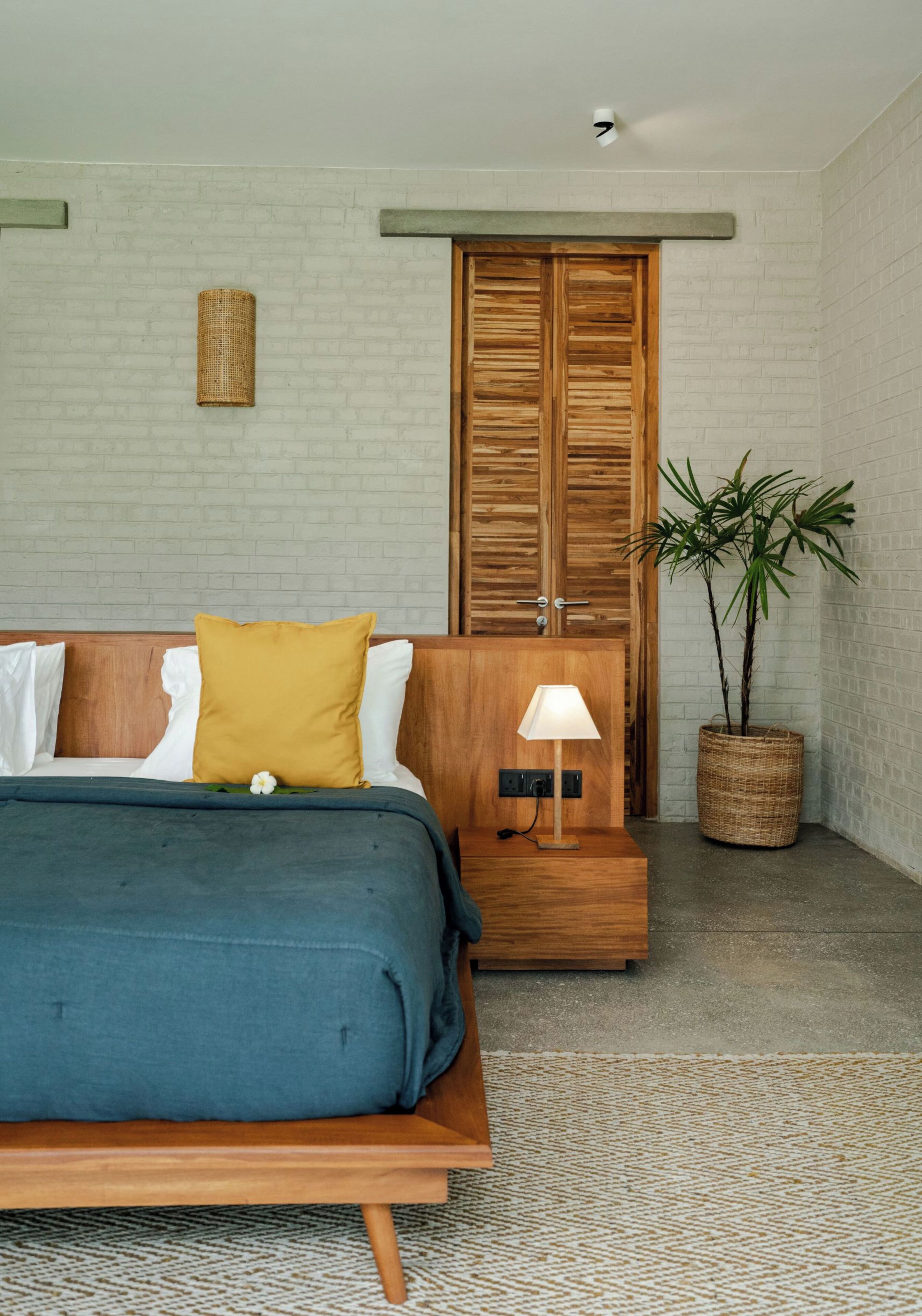 A bedroom with a bed, wooden wardrobe, bedside table, lamp, and a potted plant.