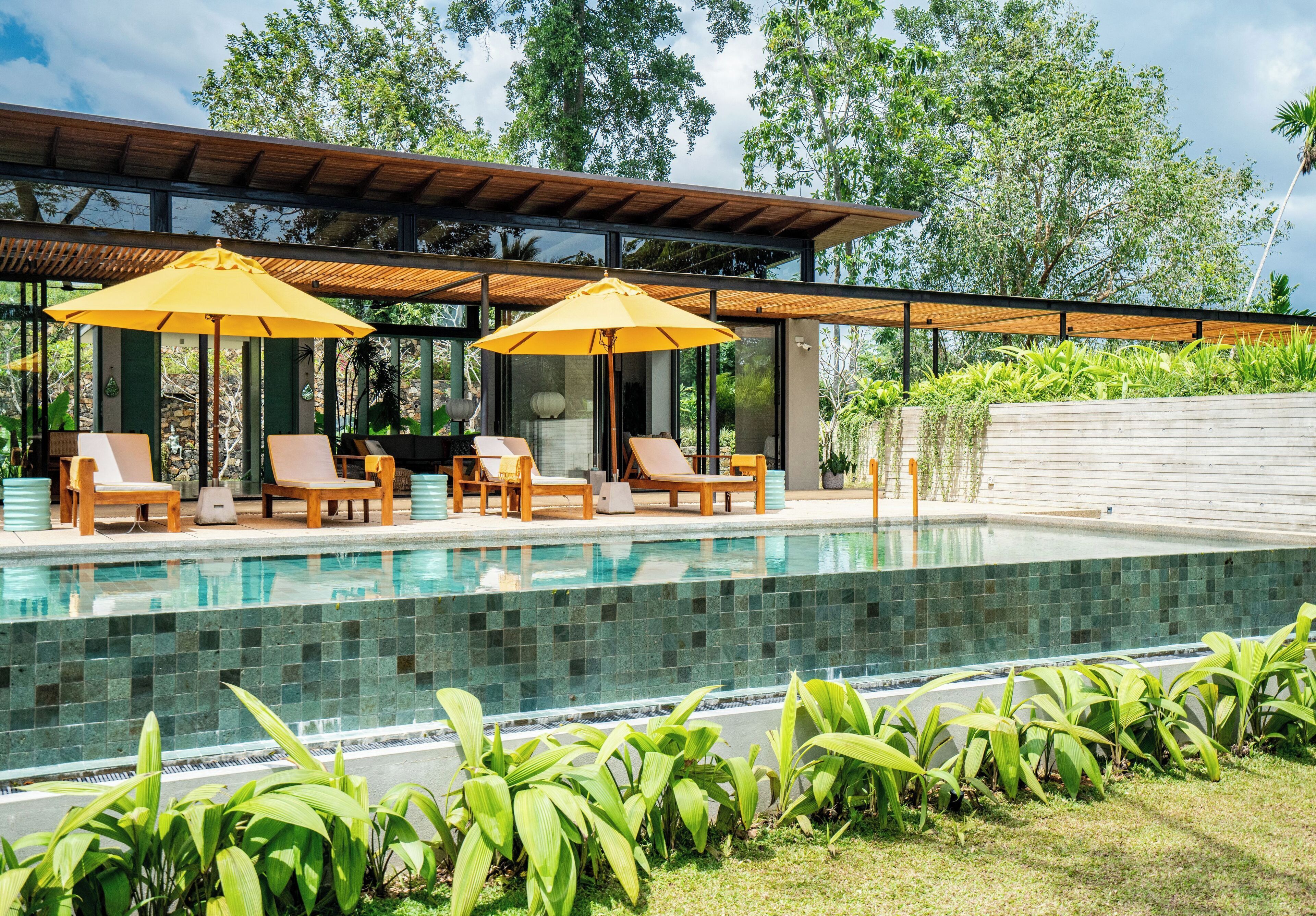 A modern outdoor pool area with lounge chairs and umbrellas, surrounded by lush greenery.