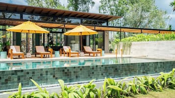 A modern outdoor pool area with lounge chairs and umbrellas, surrounded by lush greenery.