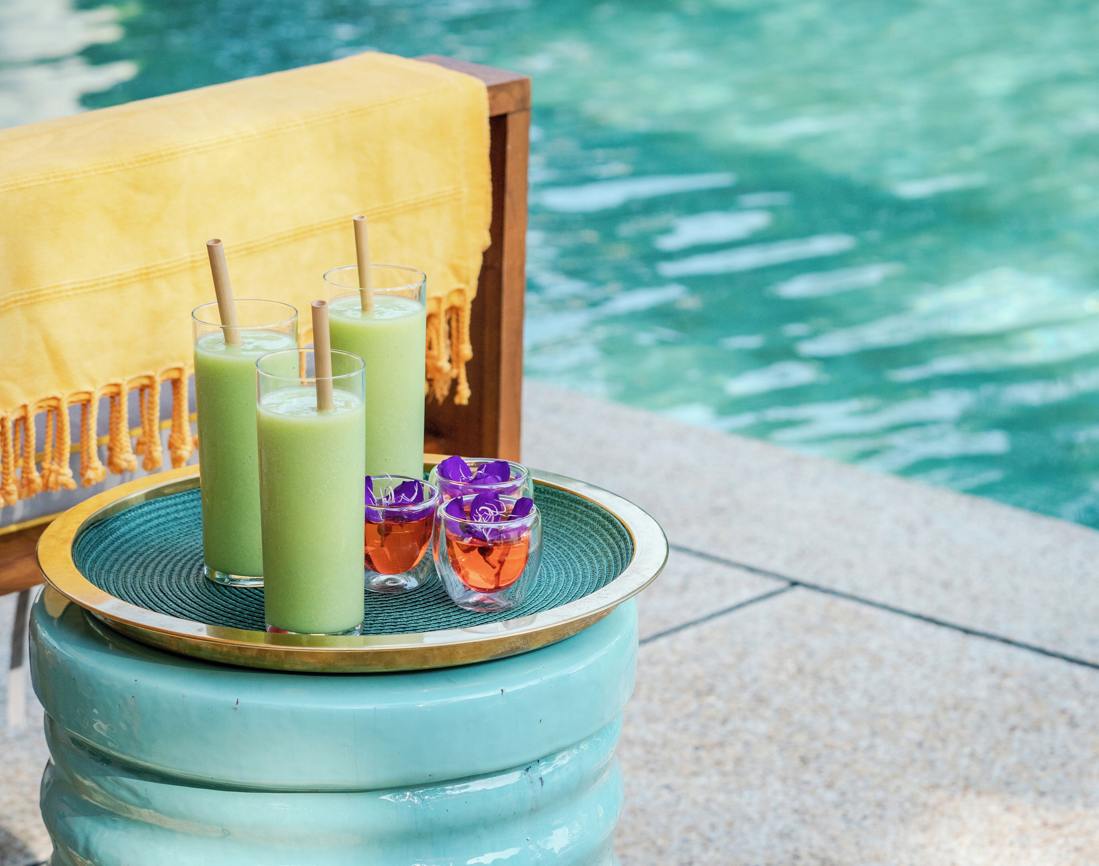 Une table avec trois boissons vertes et deux verres décorés de fleurs violettes, près d’une piscine.
