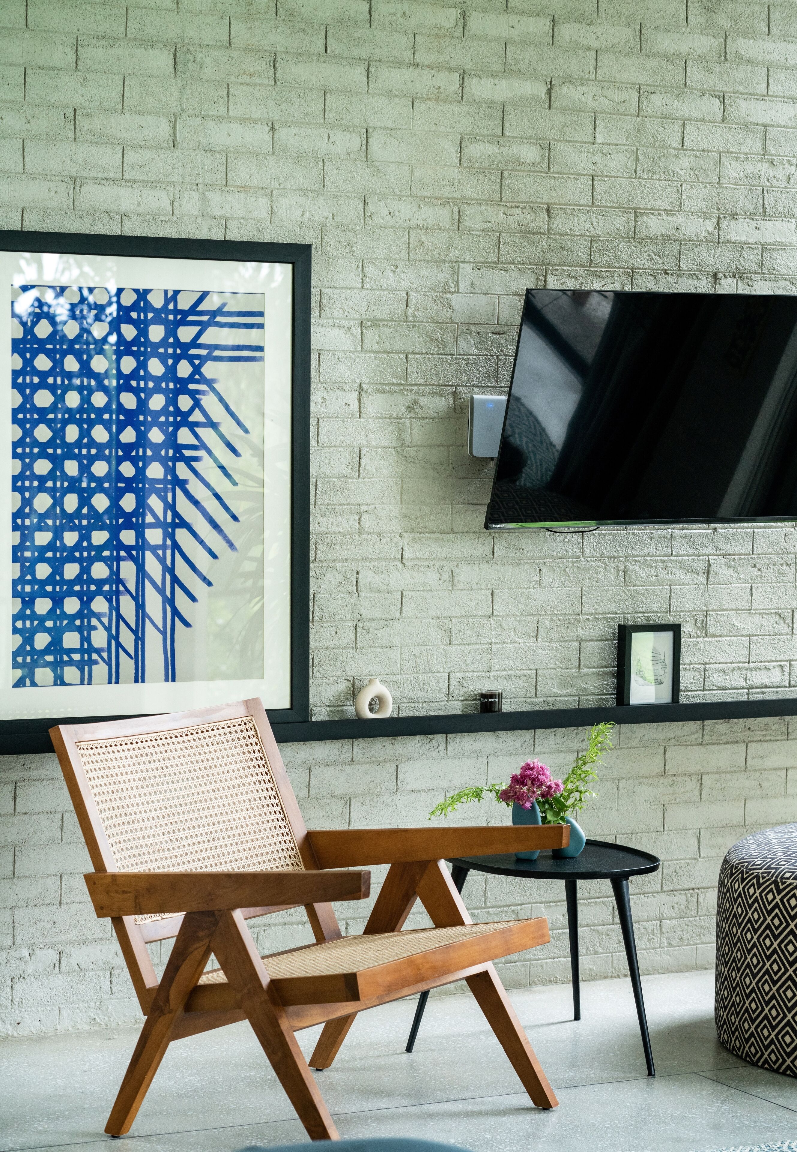Una sala de estar moderna con una pared de ladrillo, un cuadro abstracto enmarcado, una silla de madera, una mesita redonda con un jarrón y un televisor de pantalla plana montado en la pared.