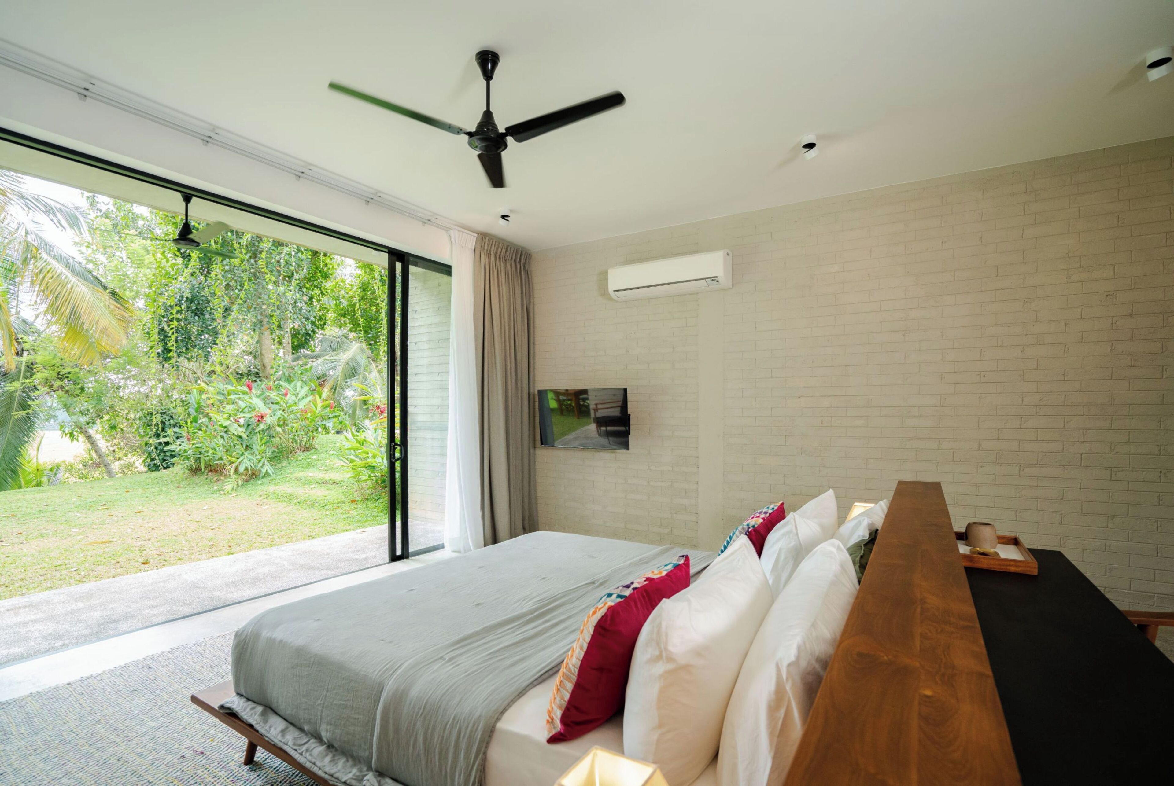 Ein Schlafzimmer mit einem großen Bett, einer hölzernen Bank, einem Deckenventilator und Blick auf einen Garten durch eine Schiebetür.