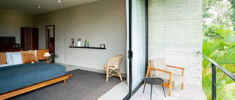 A modern bedroom with a bed, a chair, a small table, and a balcony with a view of greenery.