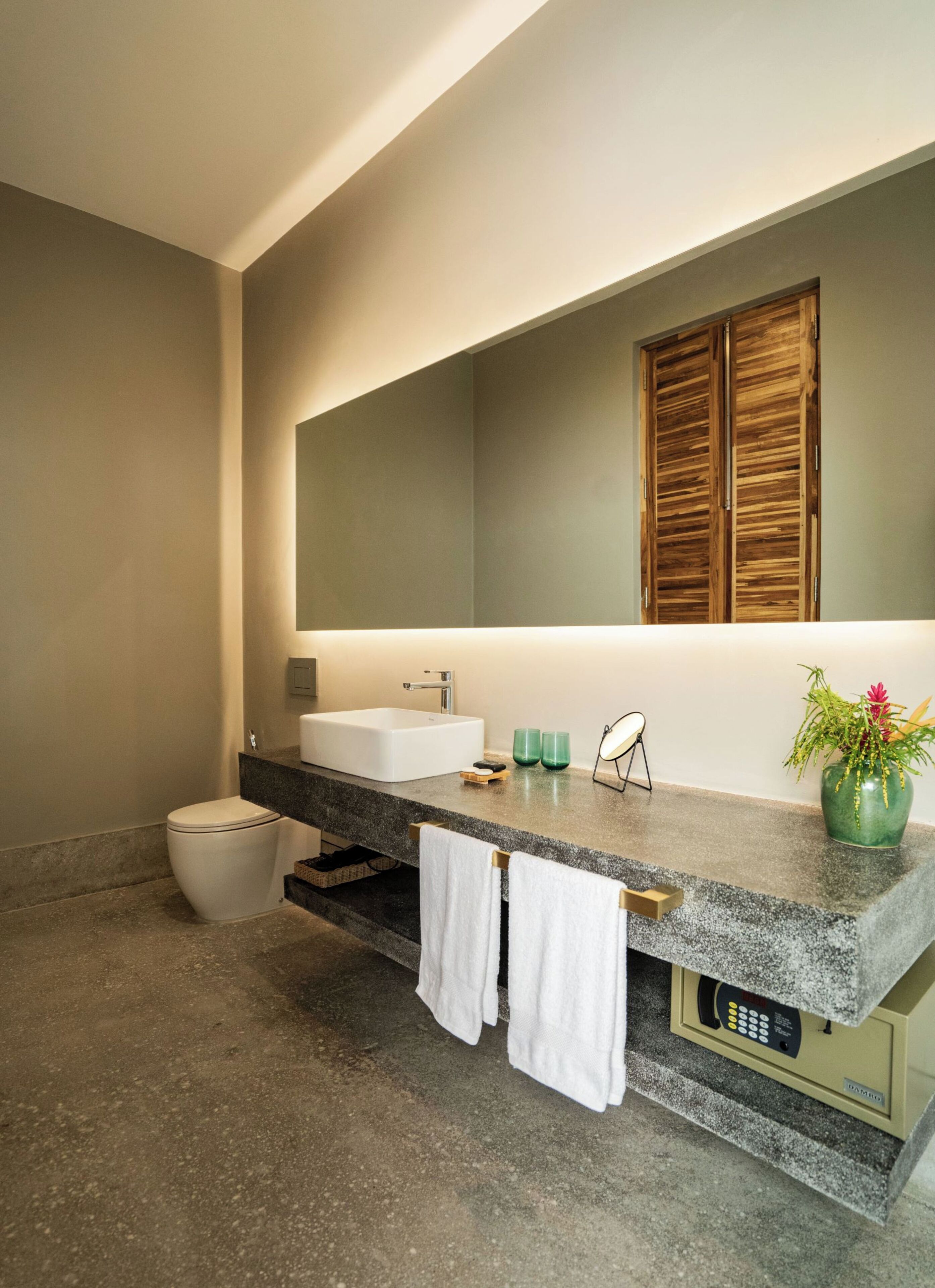 A modern bathroom with a large mirror, a white sink, and a toilet. There are two towels hanging on the towel bar, and a potted plant on the counter.