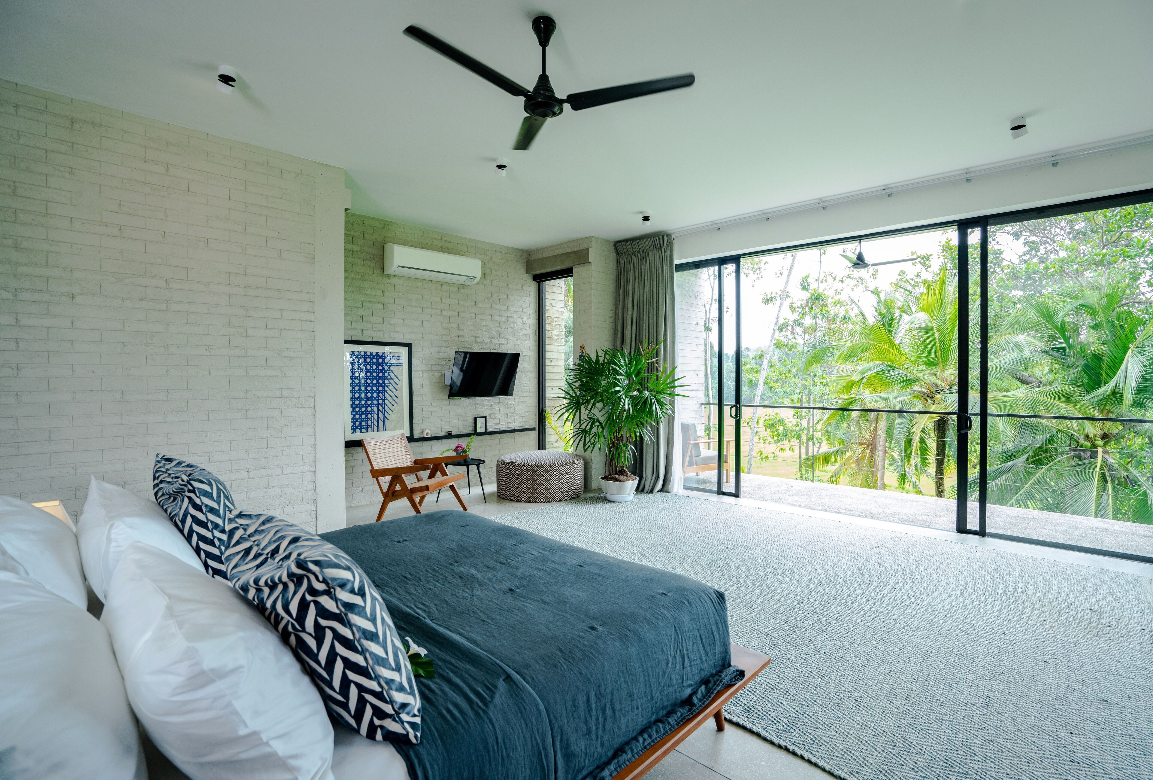 Une chambre à coucher avec un lit, un ventilateur de plafond, une télévision, une plante en pot et une vue sur la verdure grâce à de grandes fenêtres.