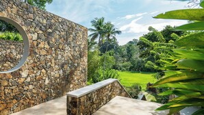 A stone wall with a circular window, a concrete ledge, and a view of a lush green landscape with palm trees. - Villa Alba (Ahangama)
