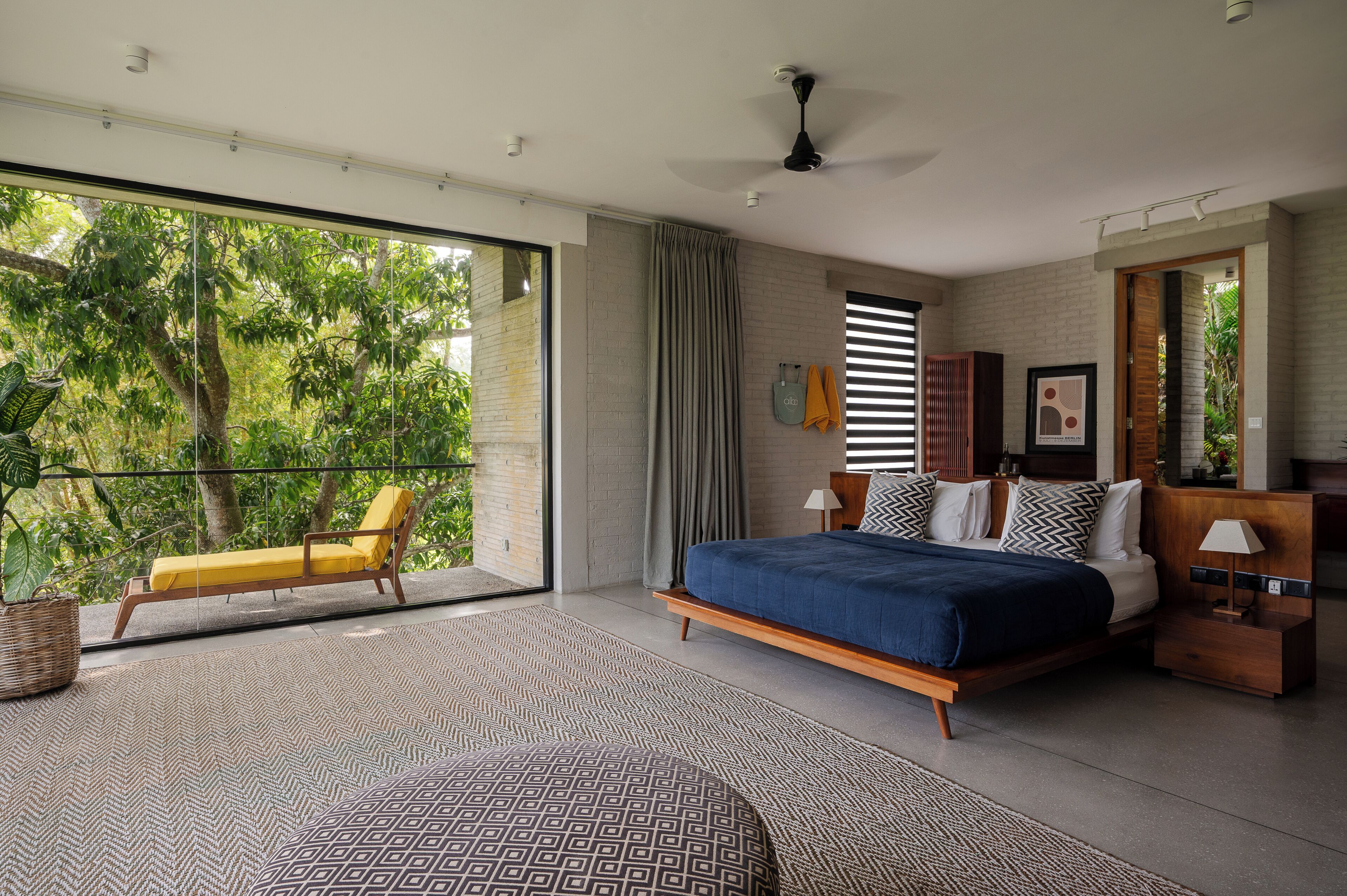 A bedroom with a large bed, a balcony with a yellow chair, and a view of trees.