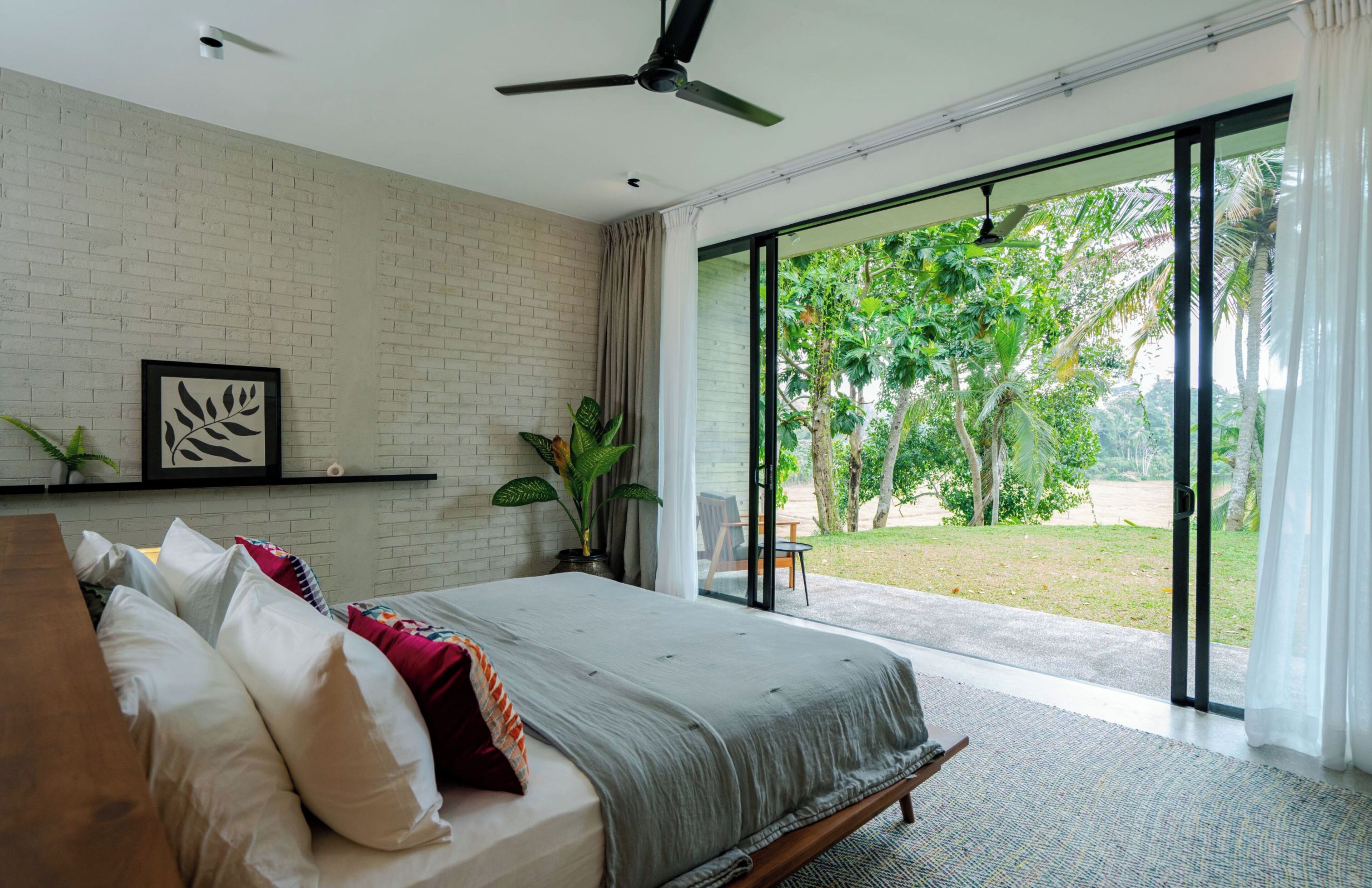 A bedroom with a large bed, a ceiling fan, a window with a view of greenery, and a brick wall.