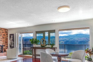 A spacious room with a large window offering a mountain view, a round table, and a seating area. - NEW LISTING Panoramic lake/mountain view, 4BR, 2Bth, Heated 20'x40' Private Pool (West Kelowna)