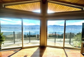 A corner room with large windows, a wooden floor, and a view of a lake and mountains. - SUNNYSIDE OASIS - Panoramic lake/mountain view, 4BR, Heated 20'x40' Private Pool (West Kelowna)