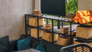 A modern living room with a large window, a flat-screen TV on a black stand, wicker baskets, and a colorful towel on a table. - Villa Kembar 2 (Canggu)