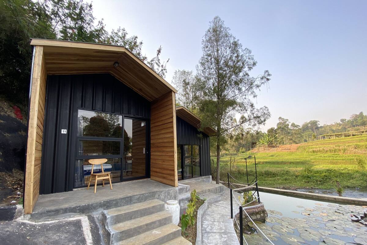 Un edificio moderno con fachada de madera y puerta de cristal, una escalera de piedra que lleva a una piscina con nenúfares y una vista de un campo verde al fondo.