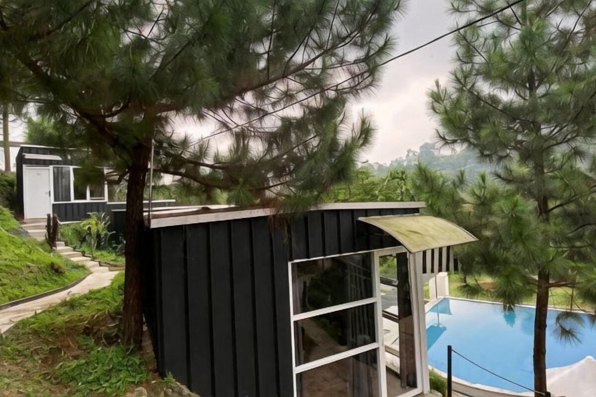 A small, dark-colored cabin with a glass door and windows, situated near a swimming pool and surrounded by trees.