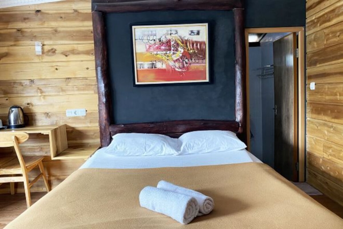 A neatly made bed with white linens and a rolled towel, a wooden desk with a kettle, and a framed abstract painting on the wall.