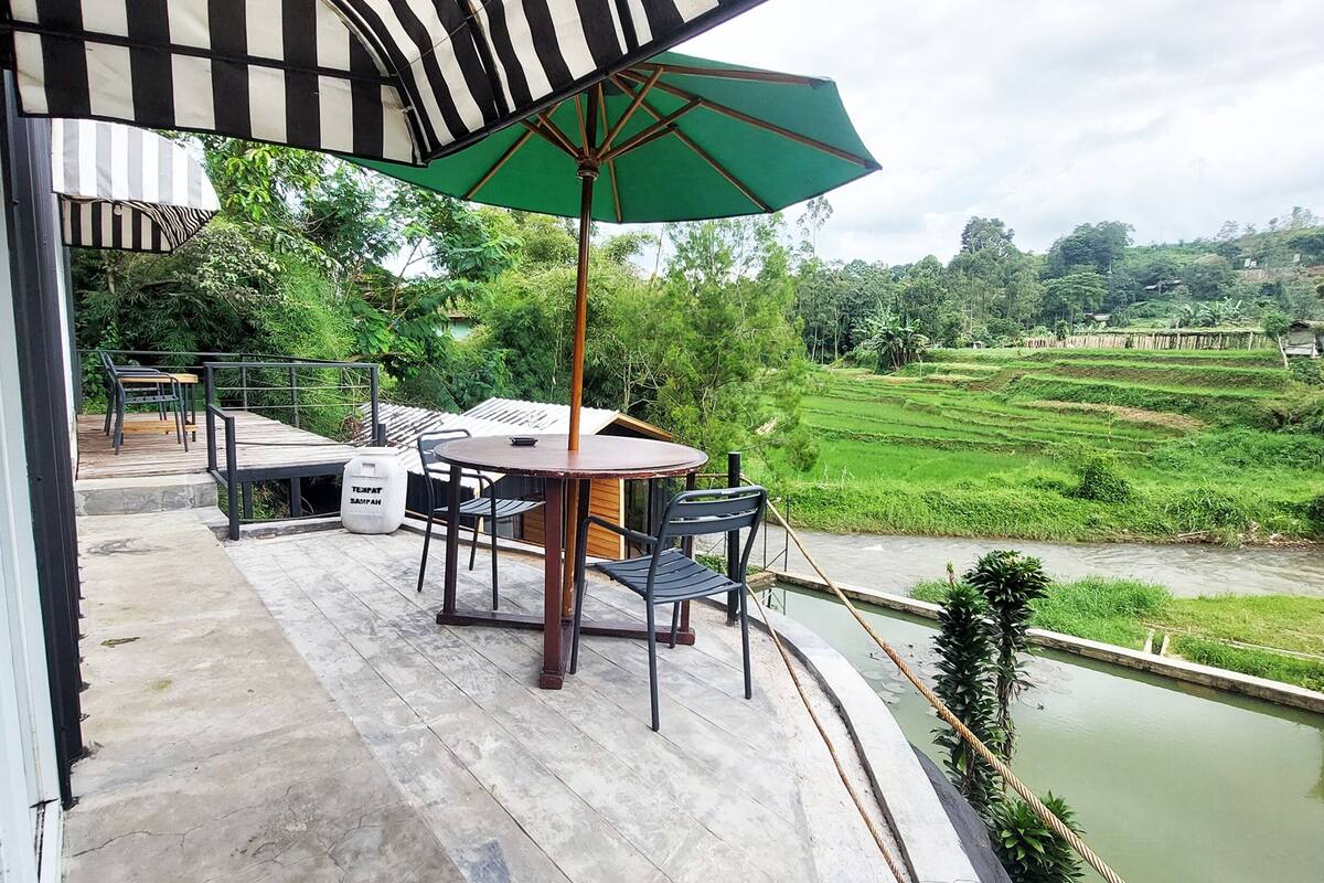Una terraza con mesa y sillas, un paraguas verde, y vista a un paisaje verde con campos escalonados.