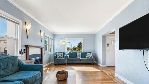 A spacious living room with a large sofa, a smaller sofa, and a coffee table. There is a fireplace, a TV mounted on the wall, and a window providing natural light.