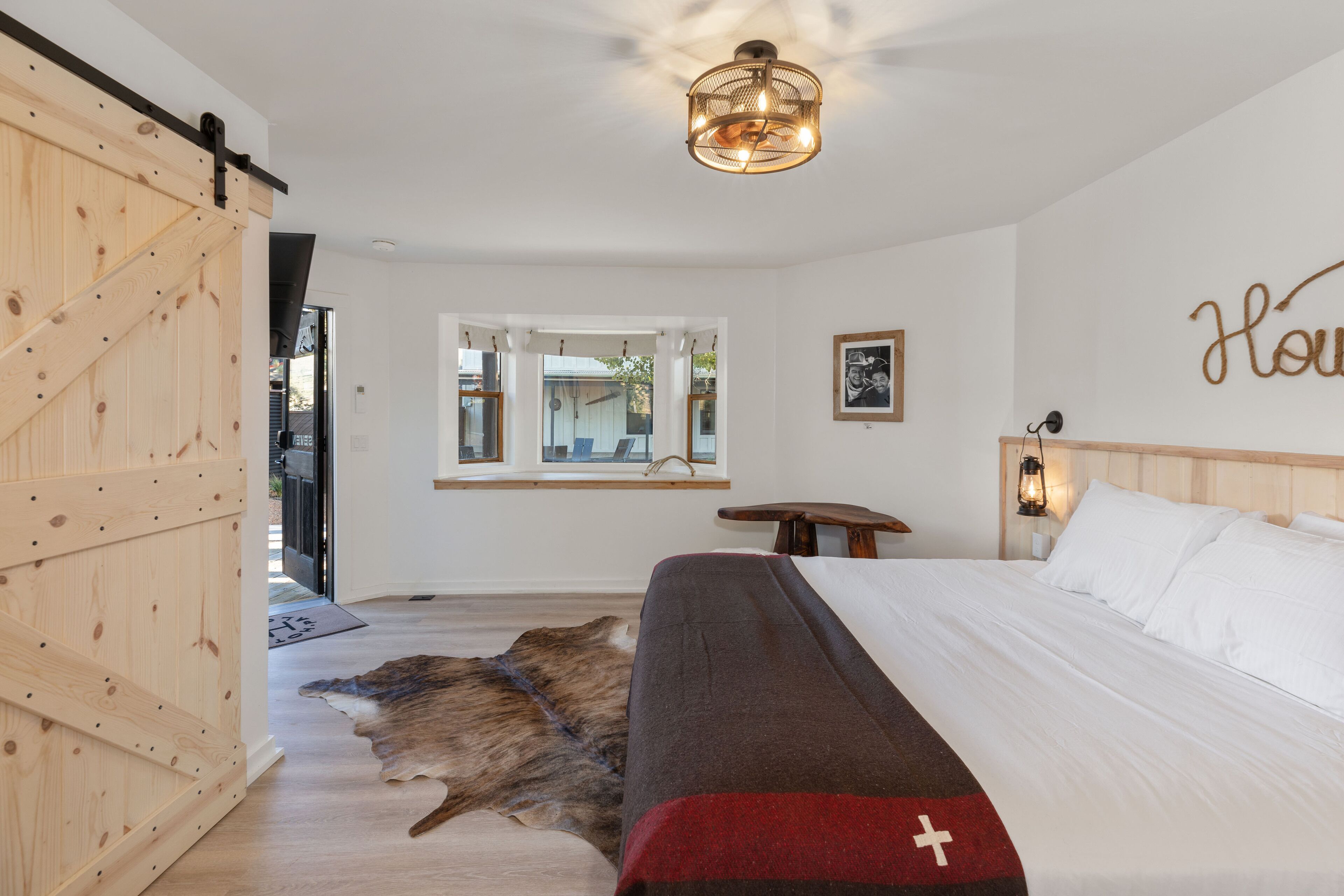 A bedroom with a bed, a wooden sliding door, a window with a view, a wall-mounted light fixture, and a framed picture.