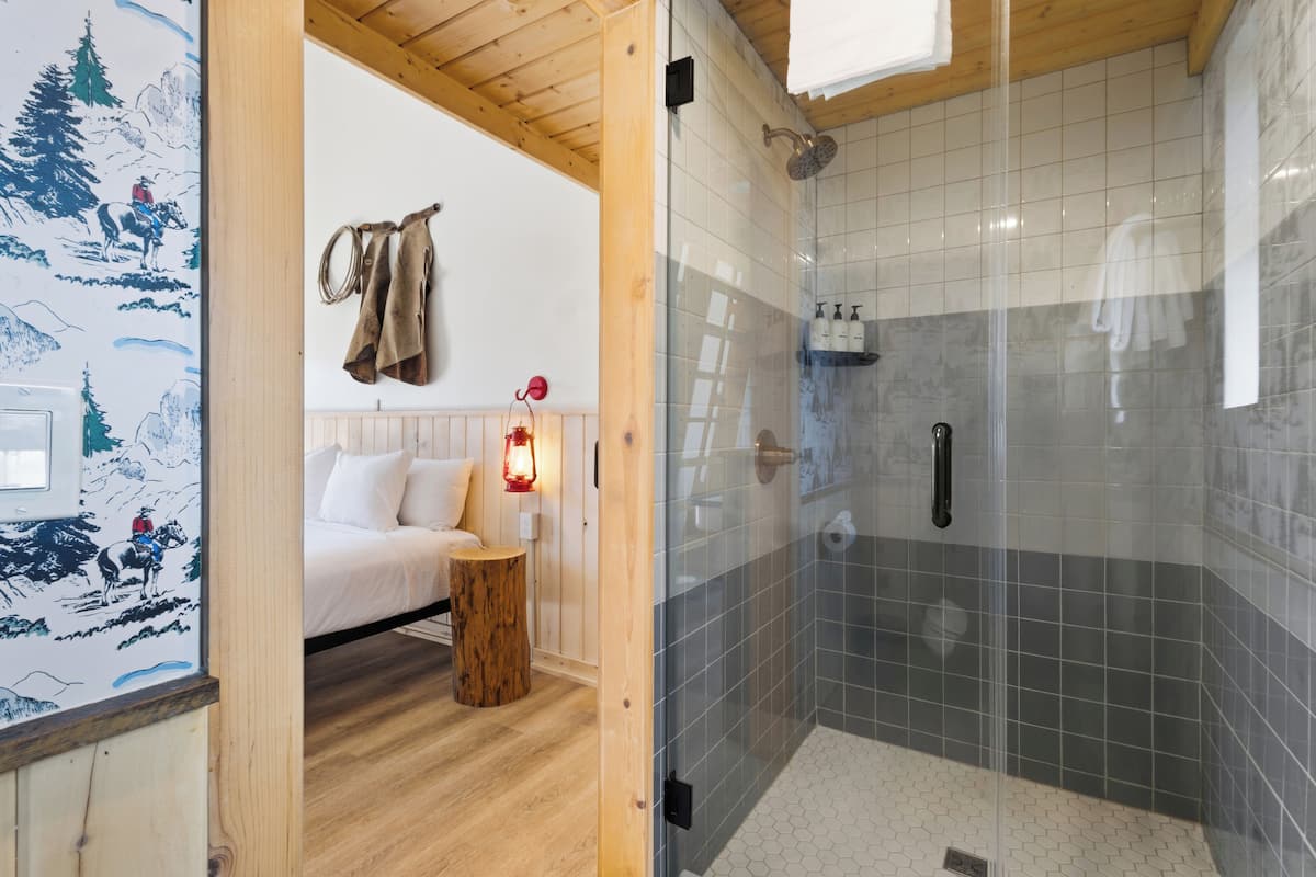 A room with a bed, a wooden side table, a glass shower enclosure, and a wall with a mountain scene.