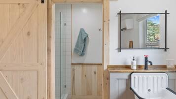 A bathroom with a wooden door, a shower with glass doors, a towel hanging on the wall, a sink with a mirror, and a window with a view of trees.