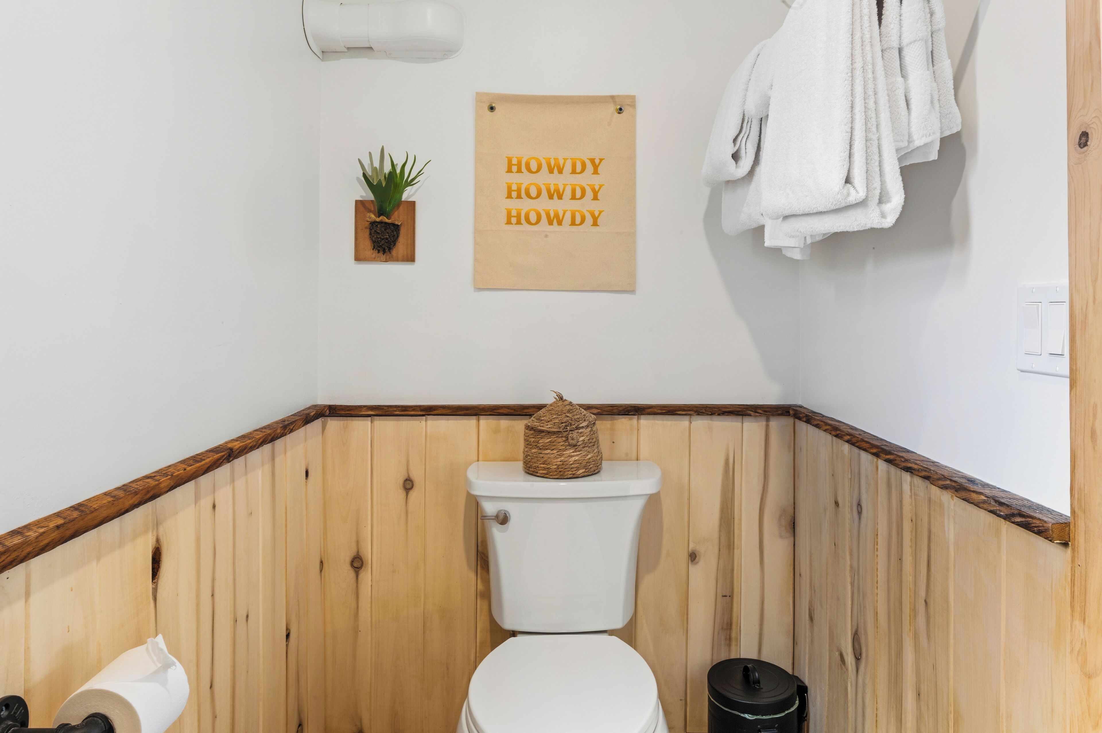 Een betegelde badkamer met een toilet, een kleine plant en een bordje met de tekst 'HOWDY HOWDY HOWDY'.