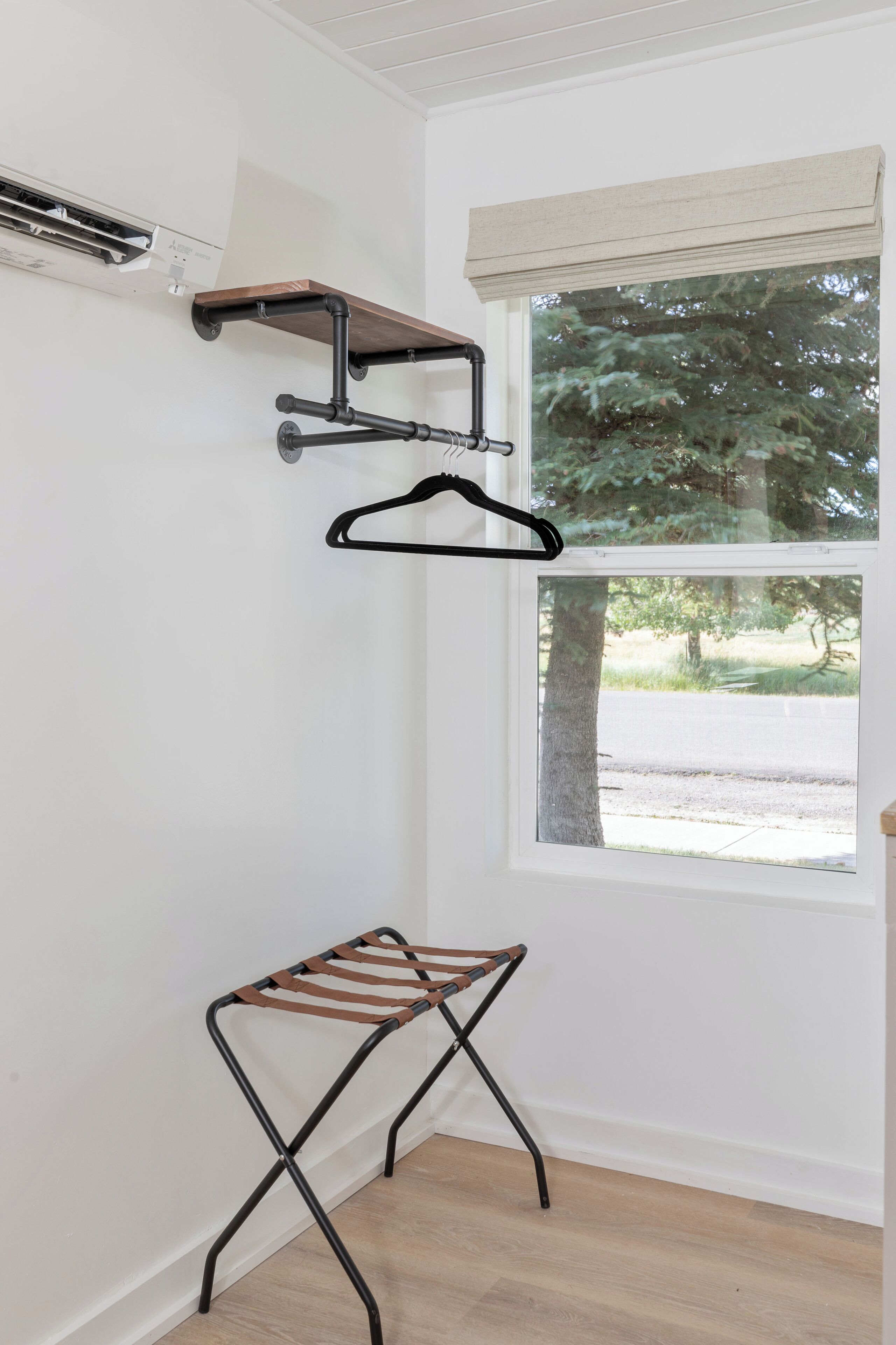 A room with a wooden floor, a window with a view of trees, a wall-mounted air conditioning unit, and a metal drying rack.