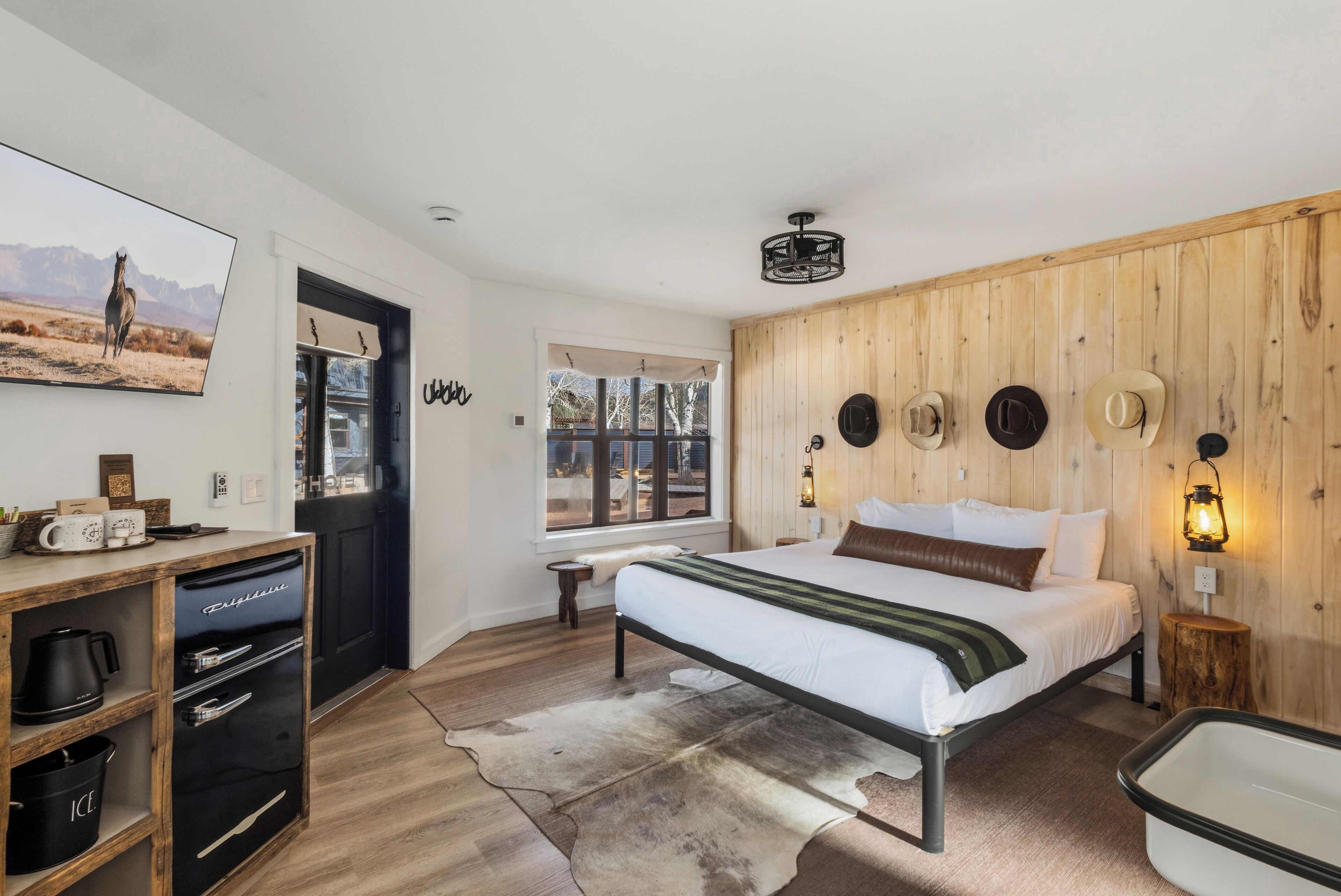 Ein Schlafzimmer mit einem Bett, einer Holzwand, einem an der Wand befestigten Hut, einer Wandleuchte, einem Wandbild, einem Schreibtisch mit Kaffeemaschine und einem Fenster.