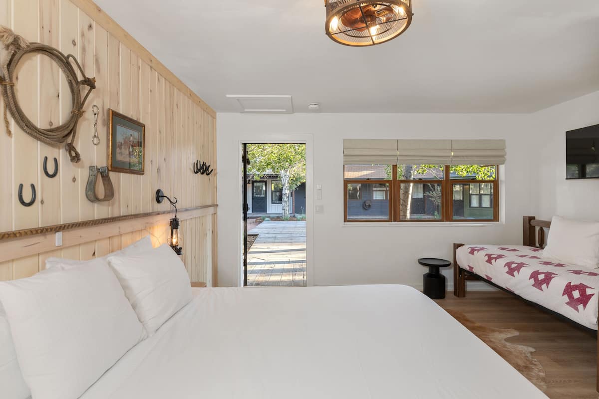 A bedroom with a large bed, a wooden headboard, a small bedside table, a bedside lamp, a window with blinds, and a door leading to an outdoor area.