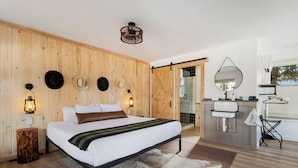 A bedroom with a wooden wall, a bed, a nightstand, a lamp, a mirror, a sink, and a view of the outdoors. - Hotel Palomino (Ridgway)