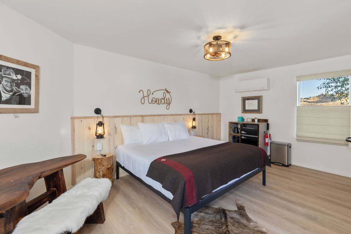 A bedroom with a wooden bed, a bench with a fur throw, a wall-mounted light, and a framed picture.