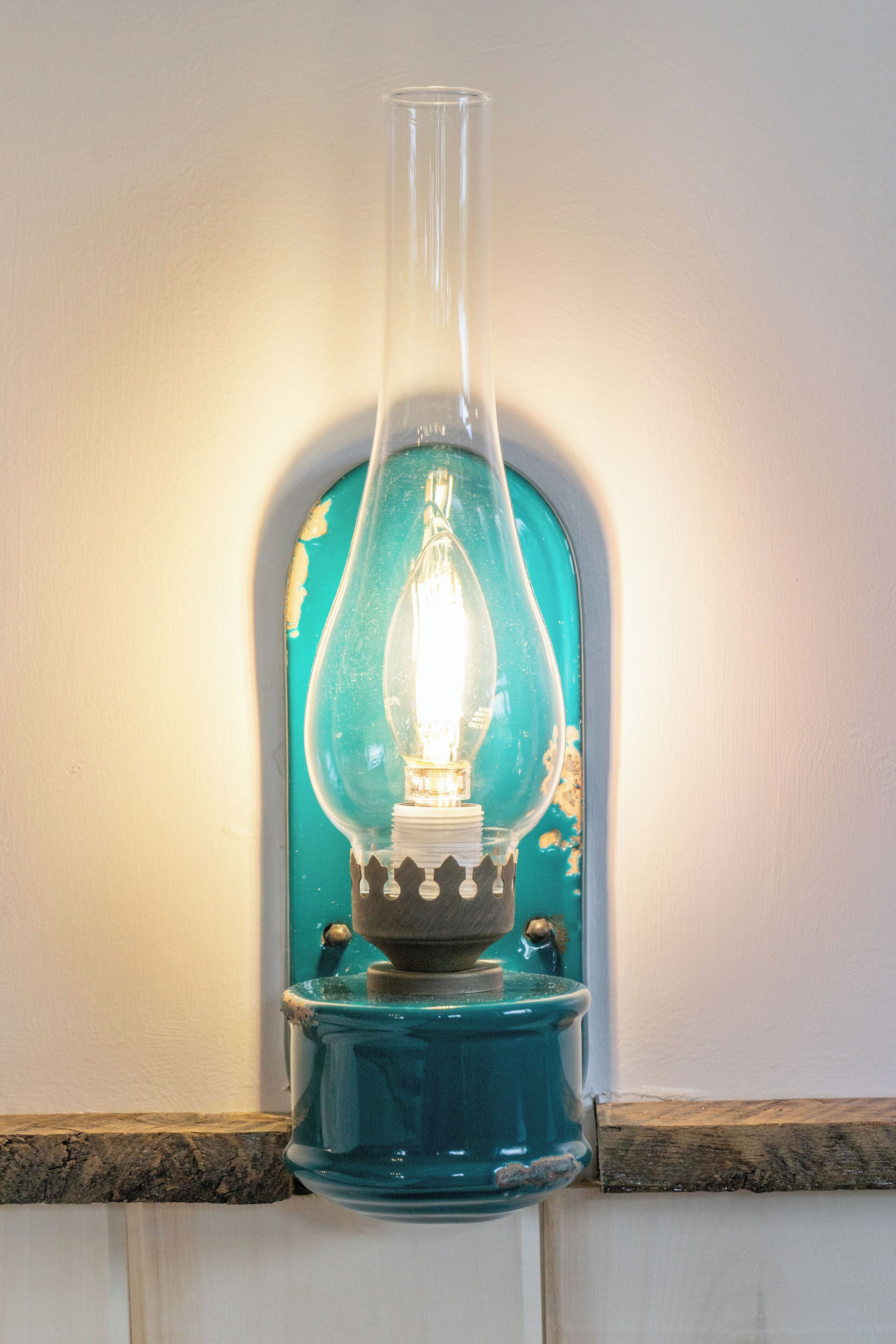 A teal-colored, vintage-style lamp with a clear glass shade and a white base, mounted on a white wall with a wooden trim.