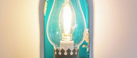 A teal-colored, vintage-style lamp with a clear glass shade and a white base, mounted on a white wall with a wooden trim.