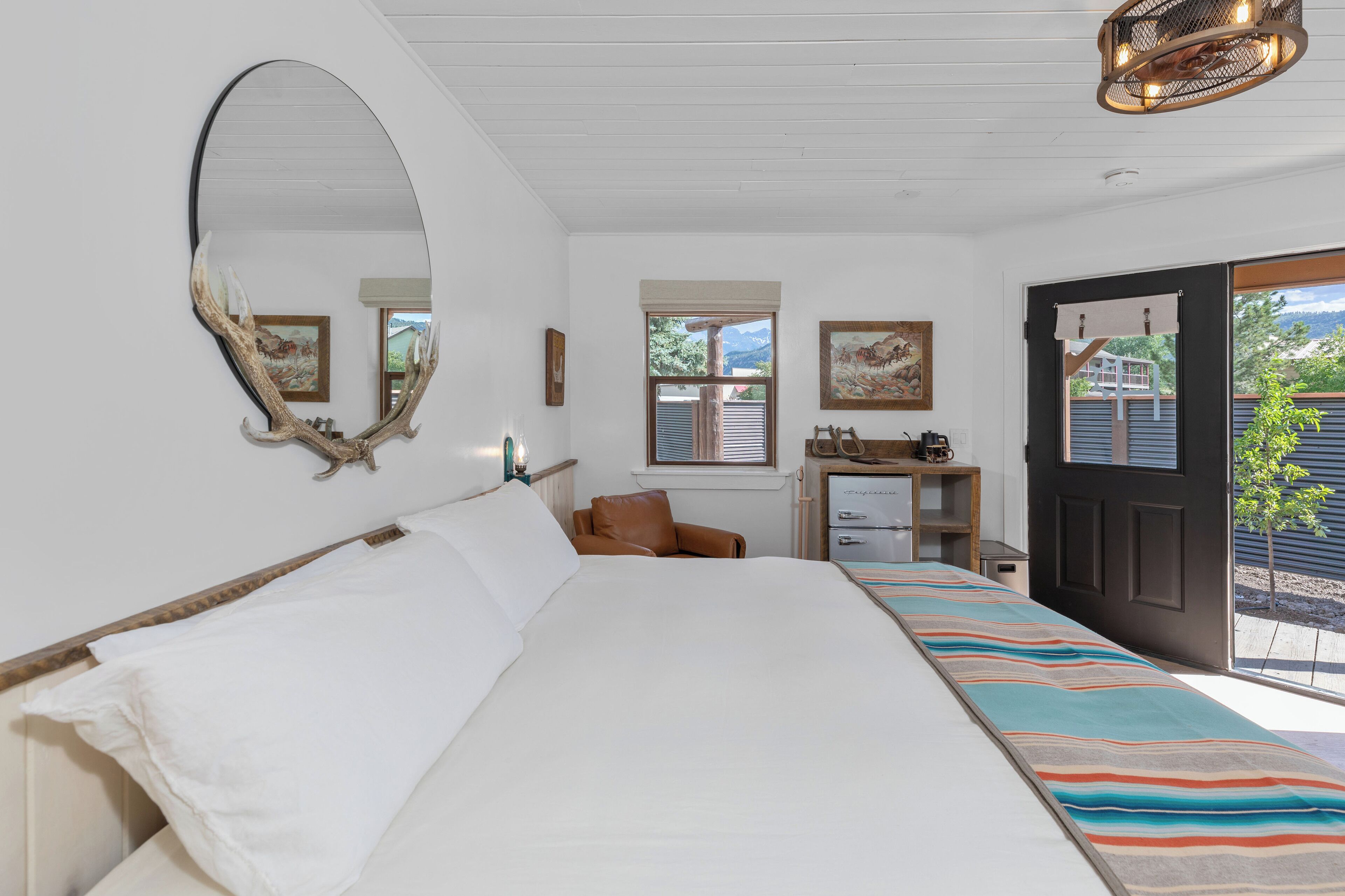 A bedroom with a large bed, a desk with a chair, a mirror, and a door leading to a balcony.