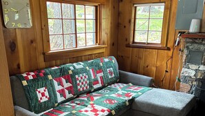 A cozy room with wooden walls, a grey sofa with a quilt, two windows, and a stone wall. - Small waterfront cabin with private dock on beautiful McGregor Lake (Marion)