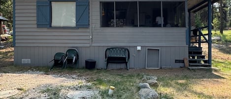 A small cabin with a porch, two green chairs, and a window overlooking a forested area.