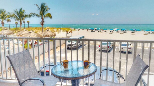A balcony with a table and chairs overlooking a beach with umbrellas and palm trees.