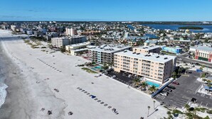An aerial view of a coastal city with a wide sandy beach, several multi-story buildings, and a marina.