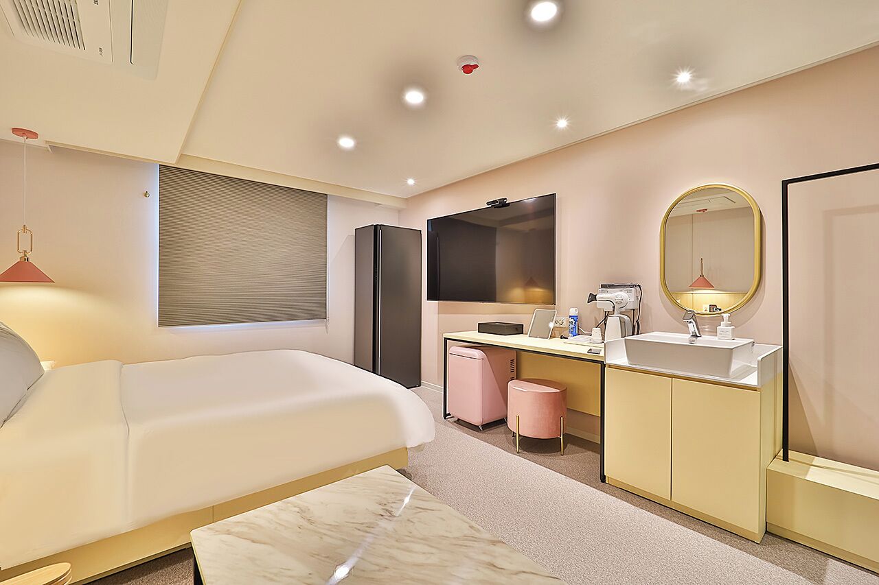 Ein modernes Hotelzimmer mit einem großen Bett, einem Schreibtisch, einem Schminktisch mit Spiegel und einem kleinen rosa Hocker.