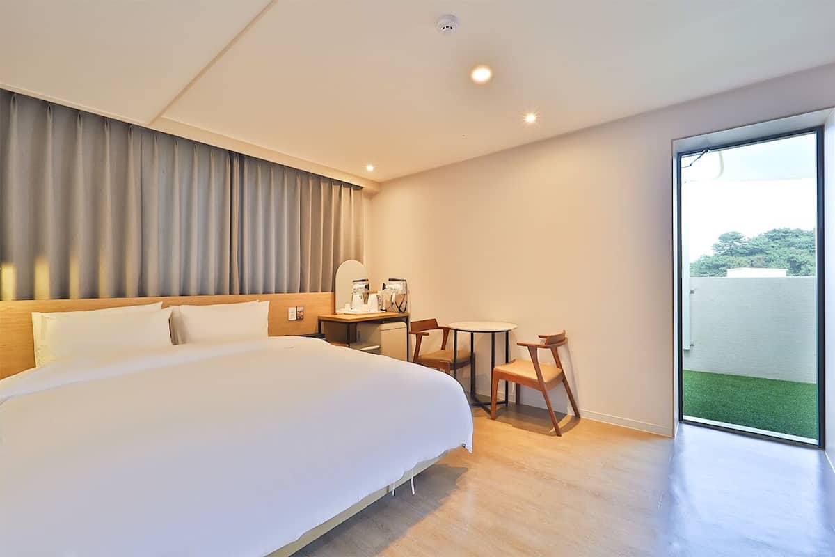 Ein modernes Hotelzimmer mit einem großen Bett, einem kleinen Schreibtisch mit Stuhl, einem runden Tisch und Blick auf eine grüne Außenanlage durch eine Glastür.