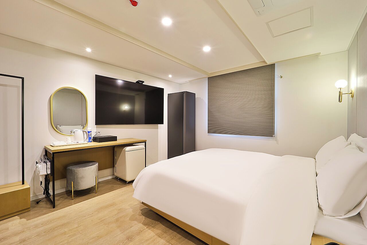 A modern hotel room with a large bed, a desk with a mirror, a wardrobe, and a window with blinds.